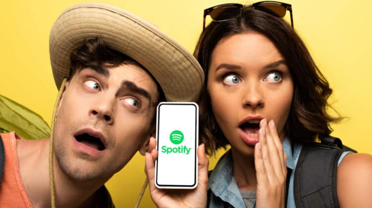 Surprised young woman showing smartphone with Spotify app while standing near shocked man on yellow background.